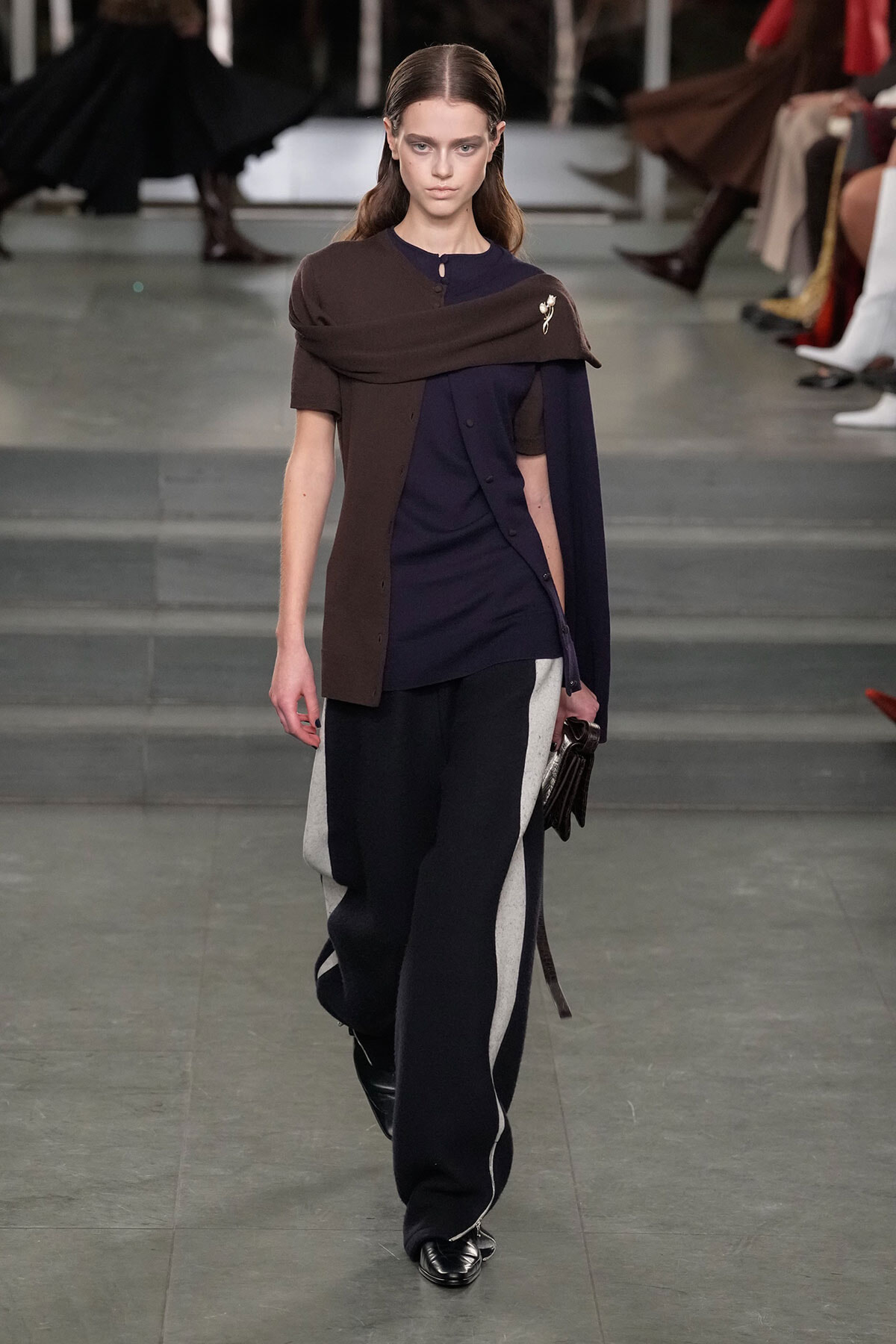 Model walking a runway in a navy tunic layered with a brown draped wrap and color-block wide-leg pants, clutch in hand