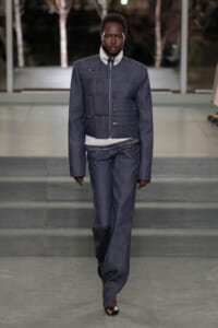 Model walks a fashion runway wearing a dark denim quilted jacket with multiple pockets and matching wide-leg jeans, layered over a light turtleneck.