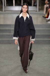 Model walks the runway in a navy sweater with a white collar, brown side-panel trousers, and a black handbag.