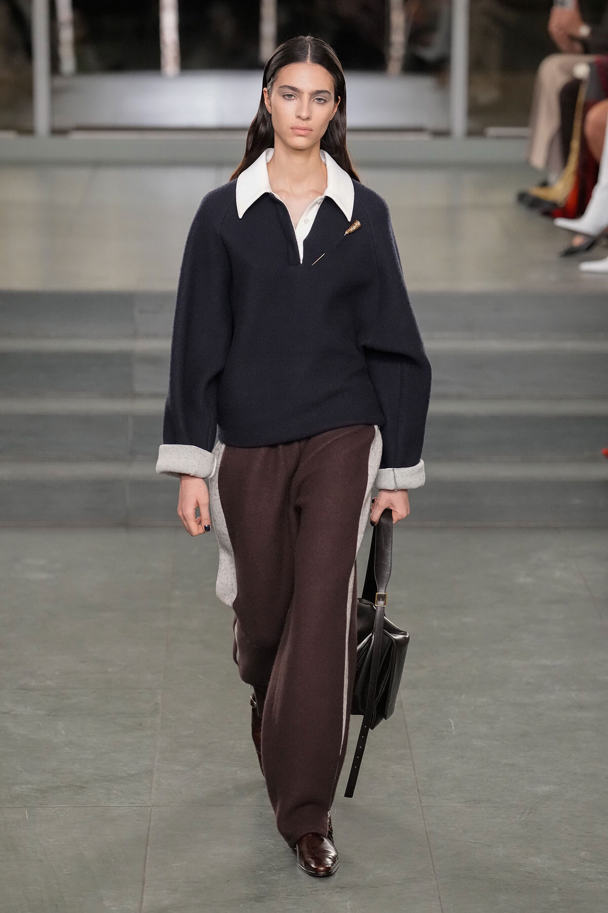 Model walks the runway in a navy sweater with a white collar, brown side-panel trousers, and a black handbag.