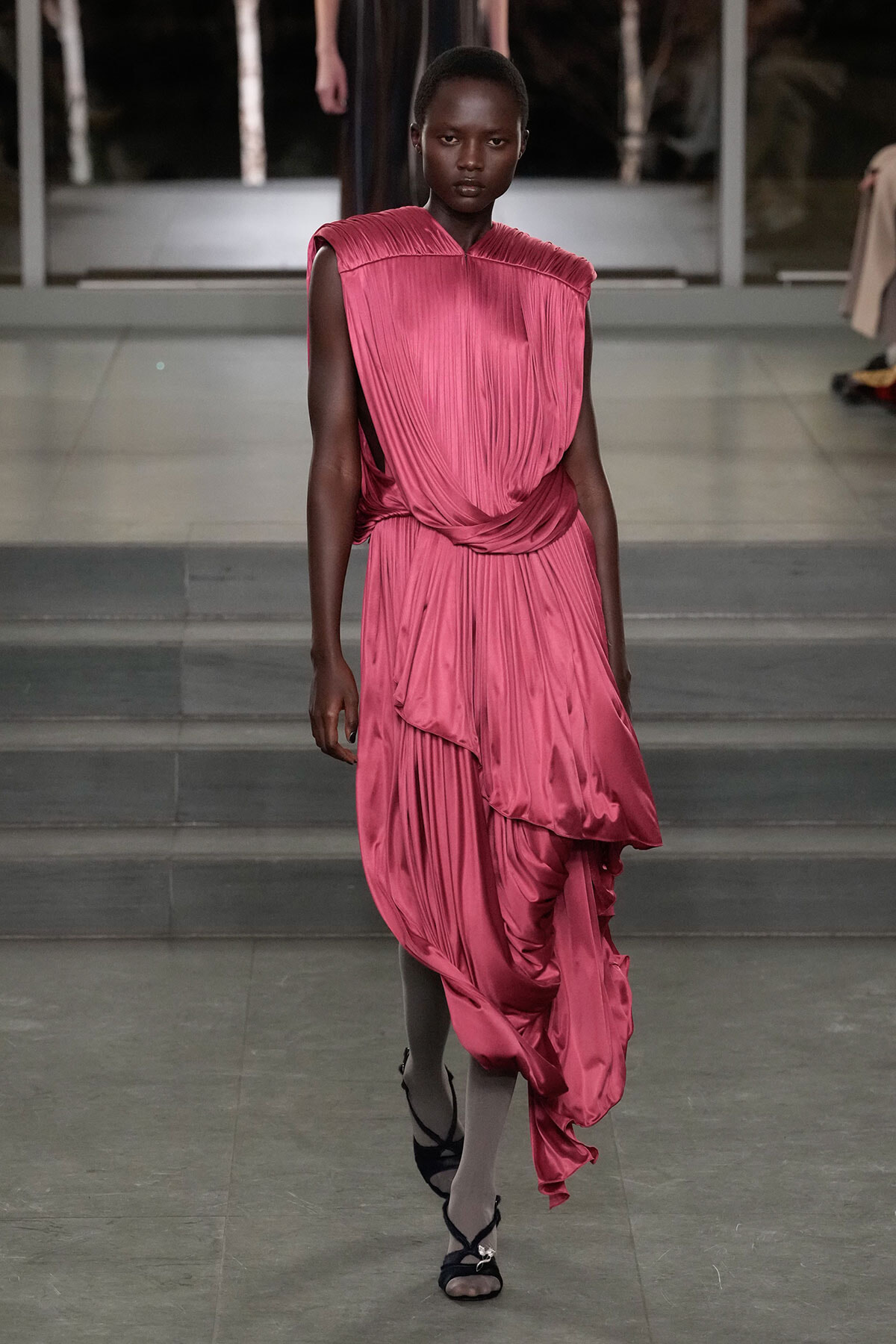 Model walks the runway in a pink, draped, pleated gown with a twisted waist.