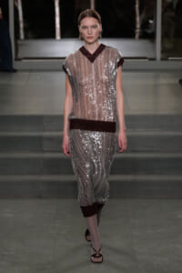 Model walks a silver sequin dress with burgundy trim along the neckline, waist, and cuffs, down a runway.