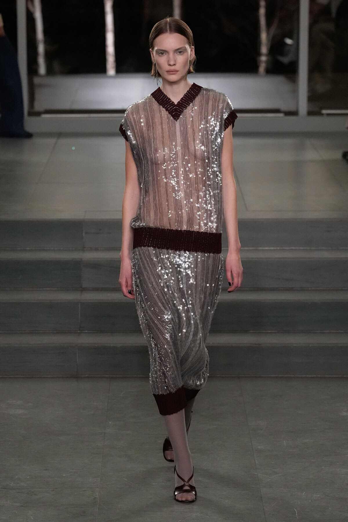 Model walks a silver sequin dress with burgundy trim along the neckline, waist, and cuffs, down a runway.