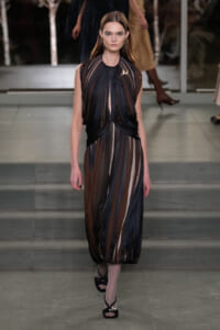 Model walks a sheer, striped dress in black, brown, and beige on a fashion runway.
