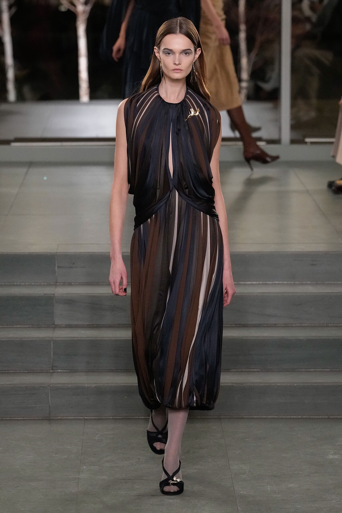 Model walks a sheer, striped dress in black, brown, and beige on a fashion runway.