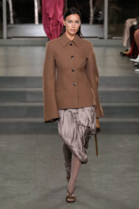 Model walking a fashion runway in a brown structured blazer with large buttons and a taupe satin draped skirt, paired with brown heels.