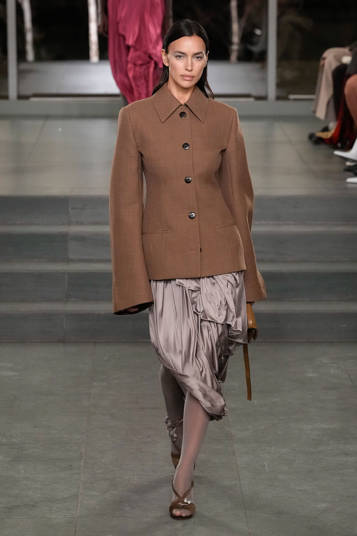 Model walking a fashion runway in a brown structured blazer with large buttons and a taupe satin draped skirt, paired with brown heels.
