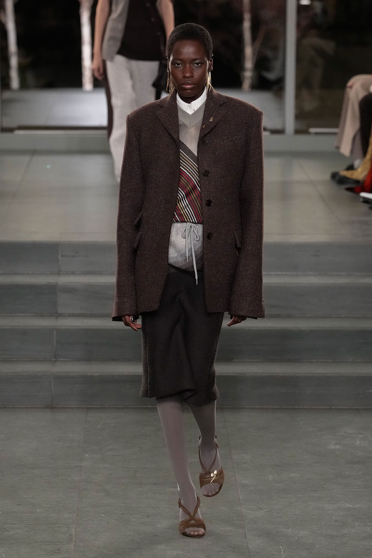 Model walks the runway wearing a brown textured wool coat over a layered knit vest and grey skirt, paired with grey tights and tan sandals.