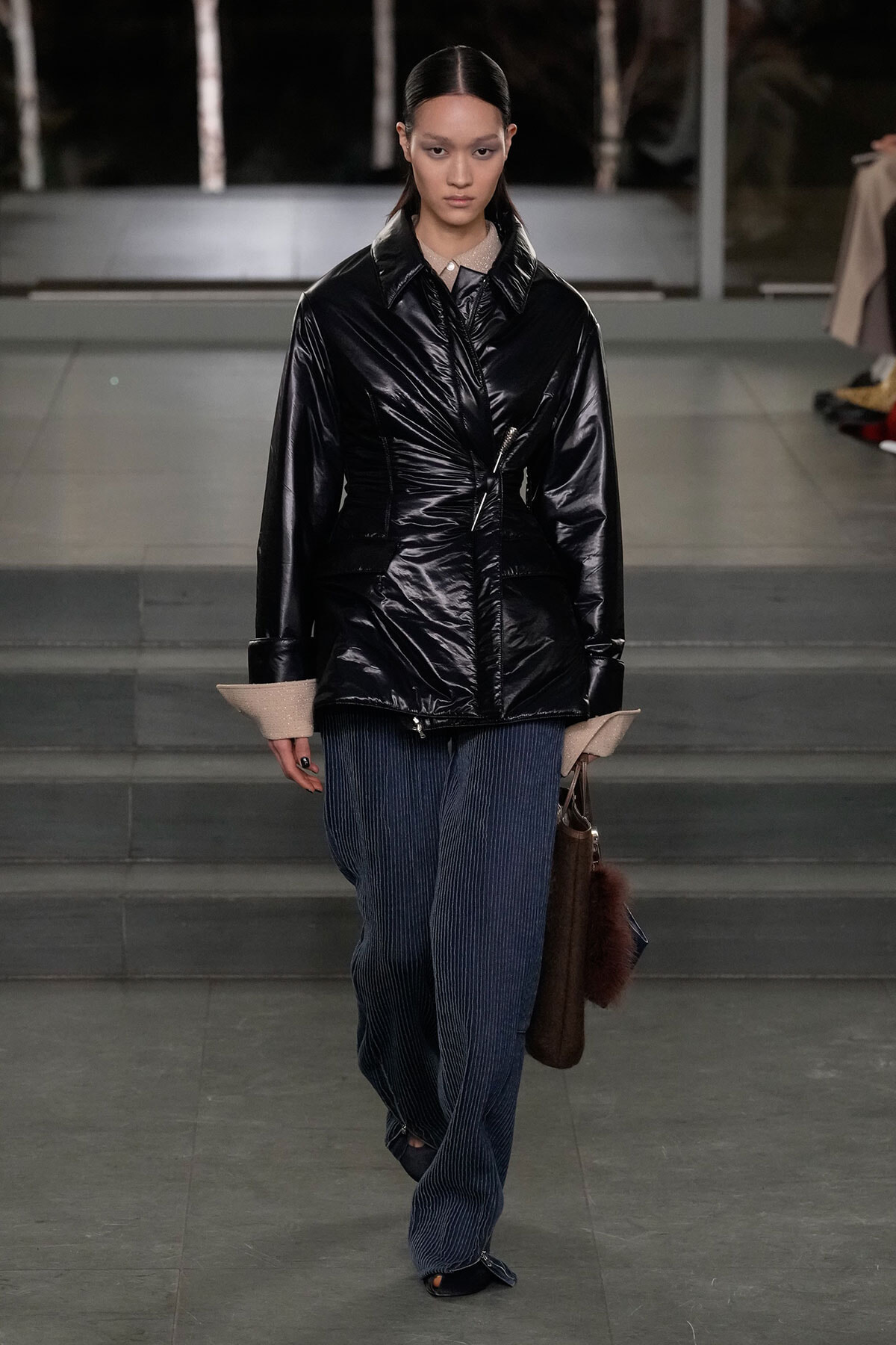 Model on a runway wearing a glossy black puffer jacket with beige cuffs and blue pinstripe wide-leg pants, carrying a brown bag.