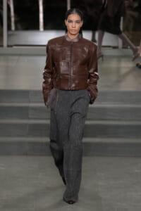 Female model strides down the runway wearing a brown leather jacket with multiple pockets and zipped front, paired with wide gray pinstripe pants.
