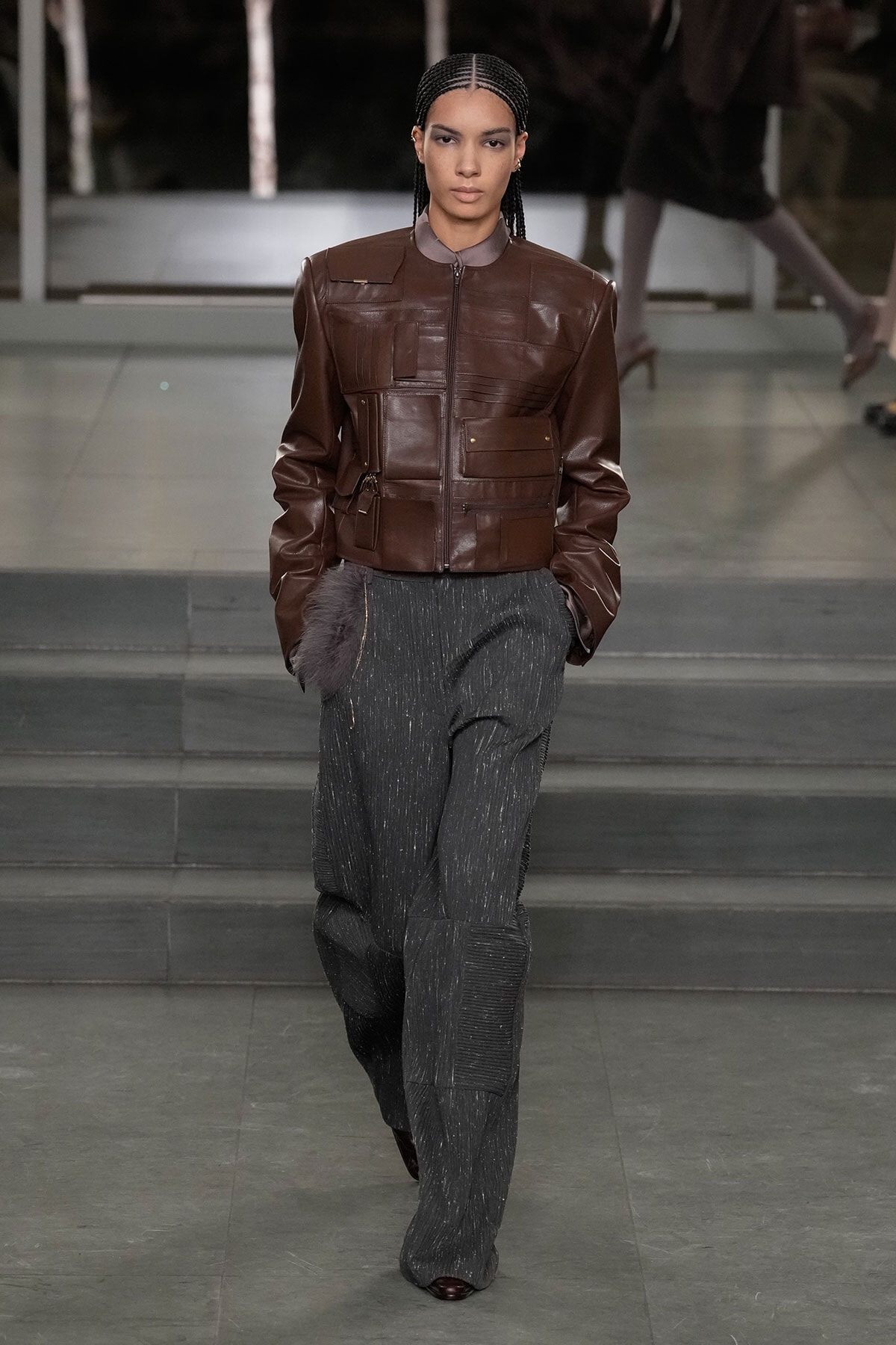 Female model strides down the runway wearing a brown leather jacket with multiple pockets and zipped front, paired with wide gray pinstripe pants.