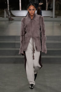 Model walking a fashion runway in a lavender-gray fur coat and light-gray wide-leg pants, black shoes, and braided hair.
