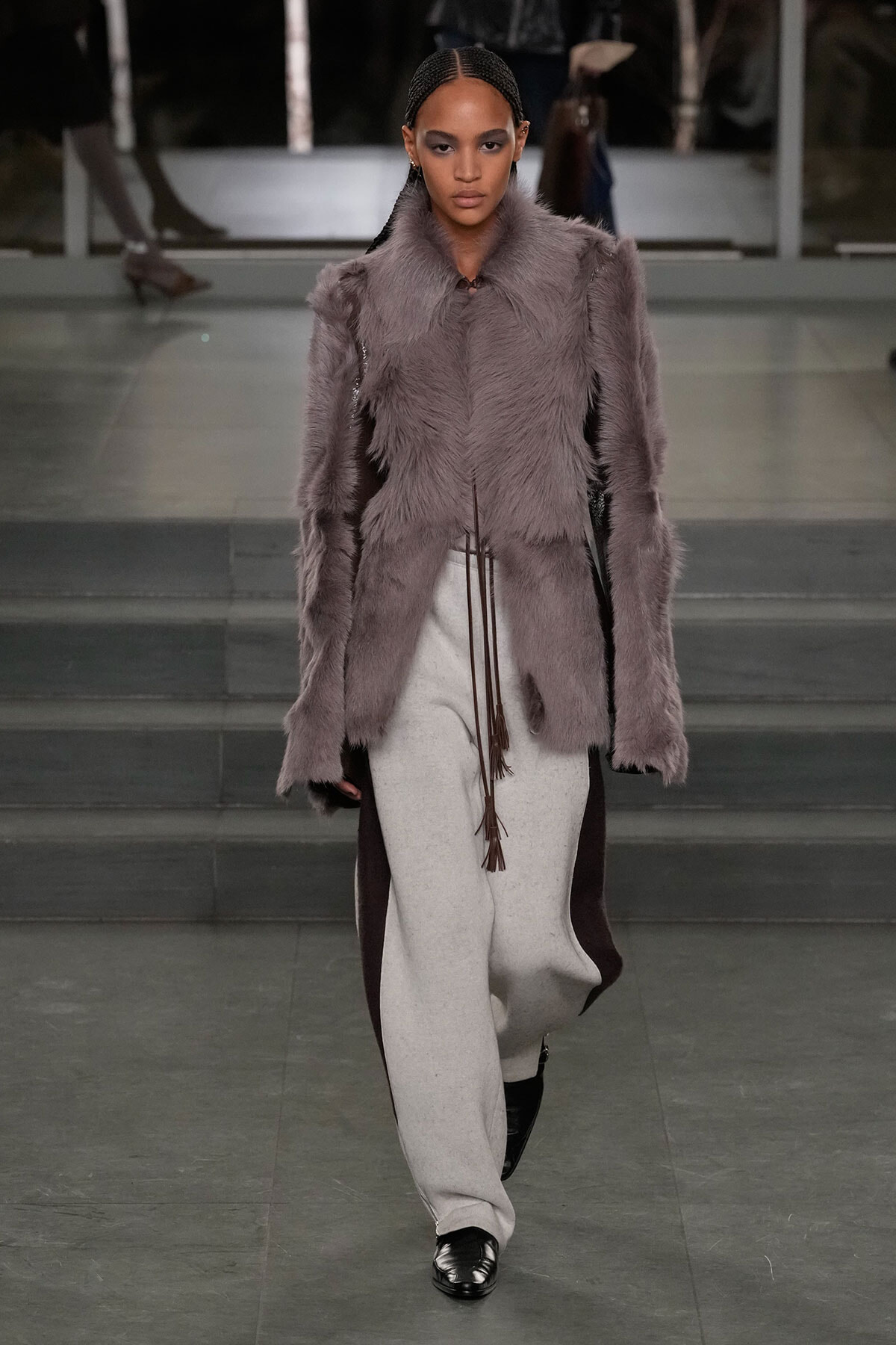 Model walking a fashion runway in a lavender-gray fur coat and light-gray wide-leg pants, black shoes, and braided hair.