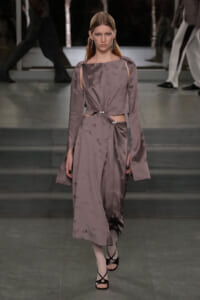 Model walks a taupe satin dress with cutouts at the waist and twisted knot detail, on a runway.