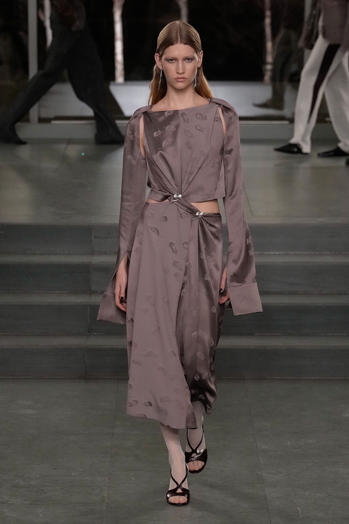 Model walks a taupe satin dress with cutouts at the waist and twisted knot detail, on a runway.