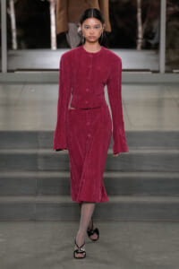Runway model wearing a magenta corduroy two‑piece: cropped button‑front jacket and matching midi skirt with side cutouts.