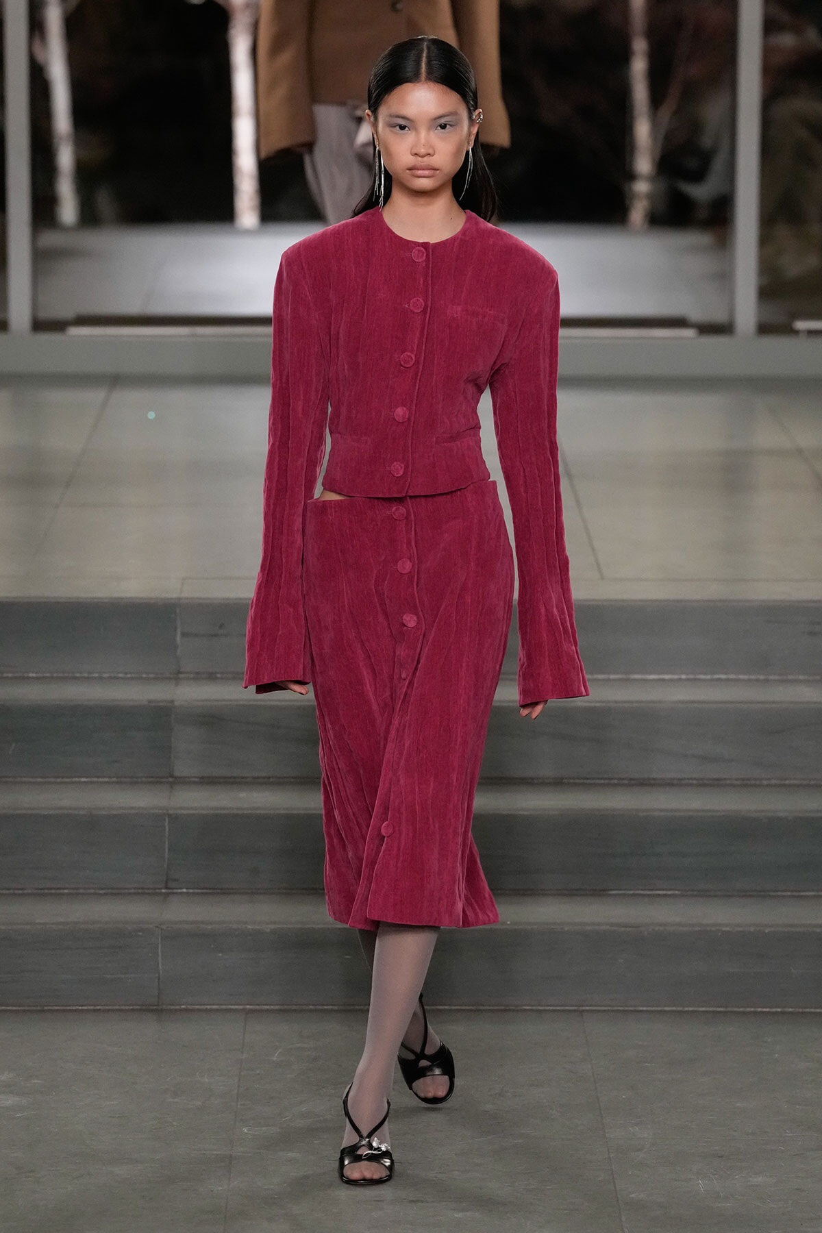 Runway model wearing a magenta corduroy two‑piece: cropped button‑front jacket and matching midi skirt with side cutouts.