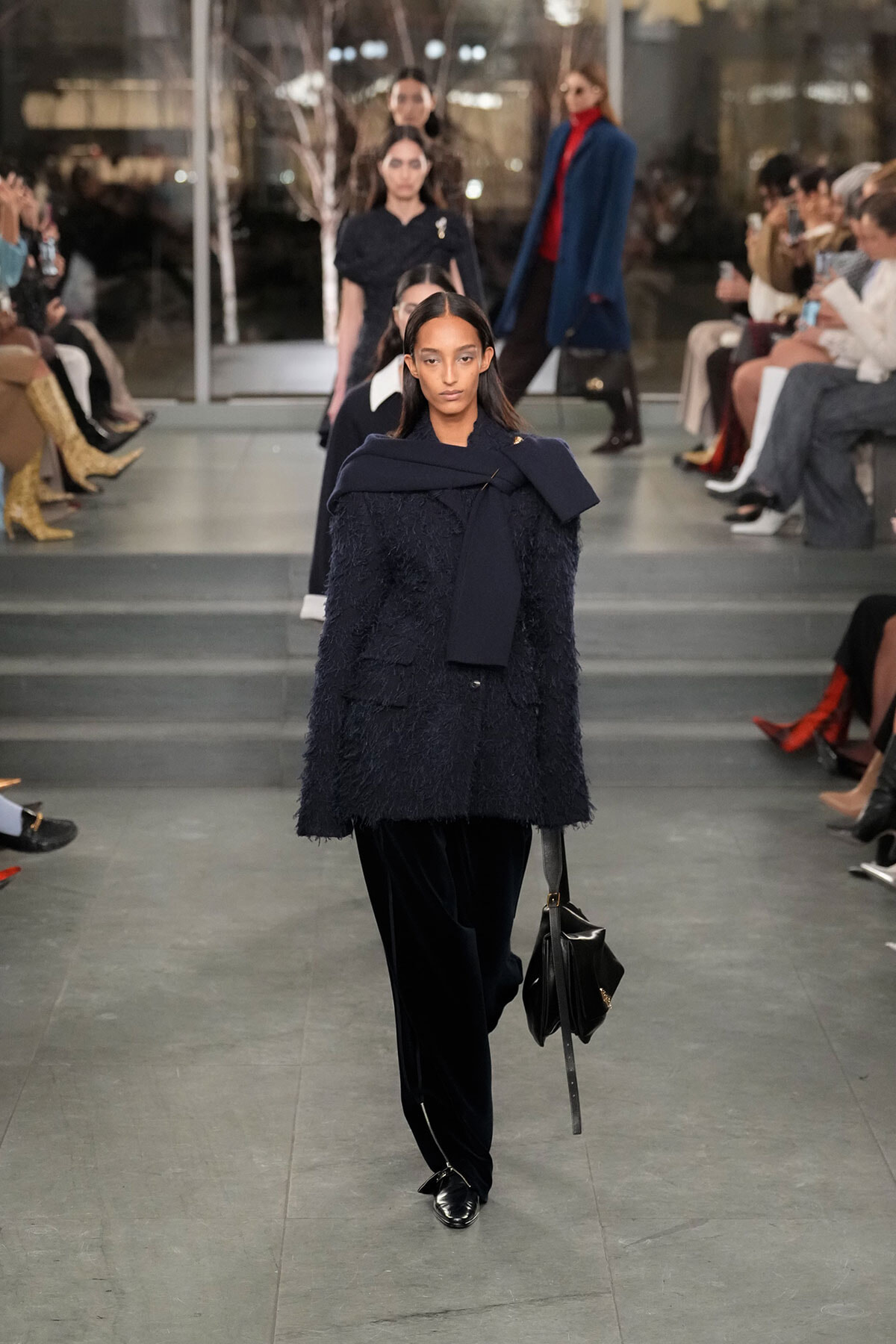 Model walks the runway in a navy textured coat with a scarf and black velvet pants, carrying a black bag.