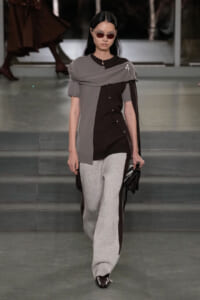 Model walking runway in a two-tone outfit: dark brown buttoned top, oversized gray shawl draped over shoulders, and wide gray speckled trousers, with red sunglasses and black shoes.