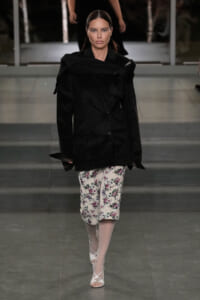 Model on a runway wearing a large black blazer over floral white culottes with pale tights and white sandals.