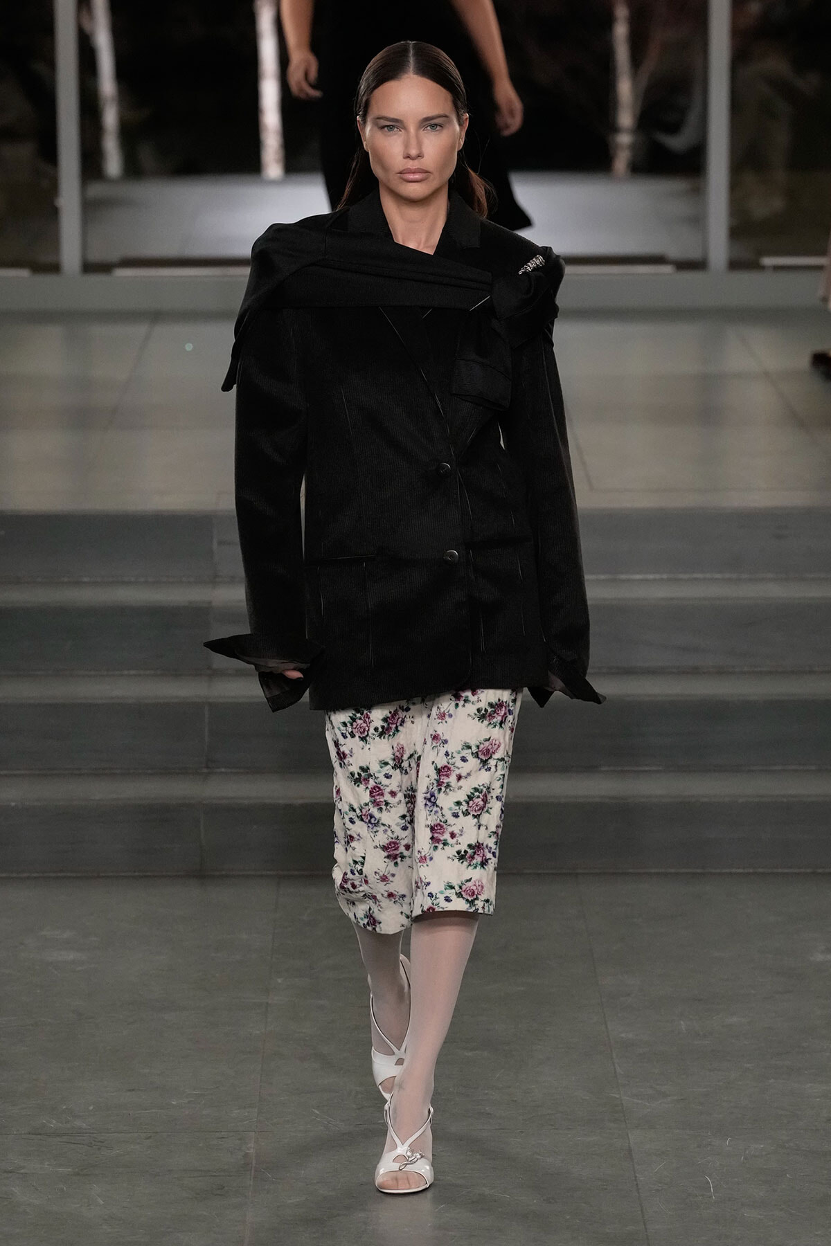 Model on a runway wearing a large black blazer over floral white culottes with pale tights and white sandals.