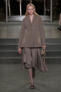 Model walks a muted taupe ensemble with an oversized blazer and flowing midi skirt on a runway, holding a small beige handbag as she strides forward.