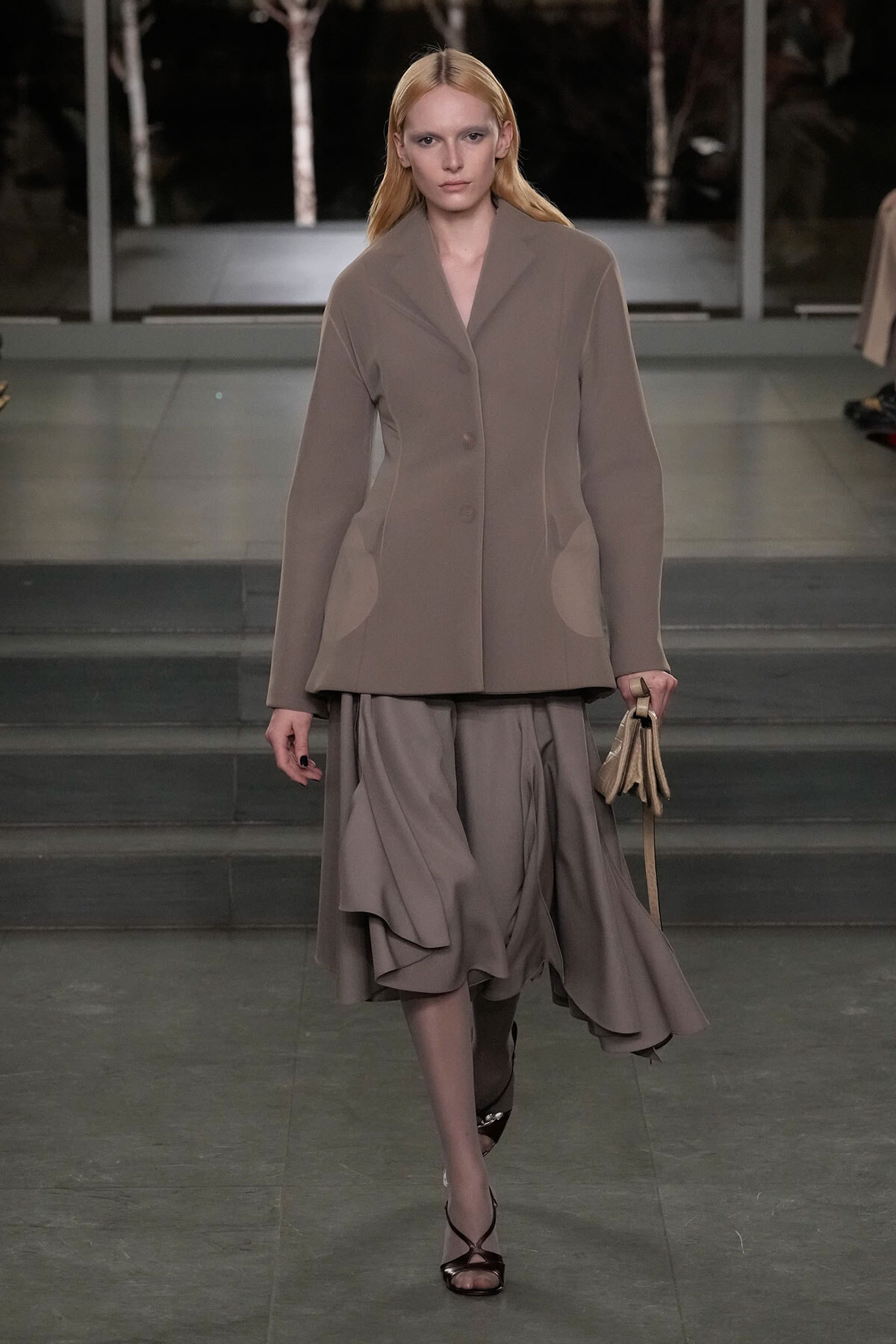 Model walks a muted taupe ensemble with an oversized blazer and flowing midi skirt on a runway, holding a small beige handbag as she strides forward.