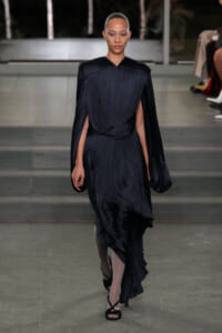 Model walks the runway in a dark navy pleated gown with cape sleeves and a draped, twisted waist.