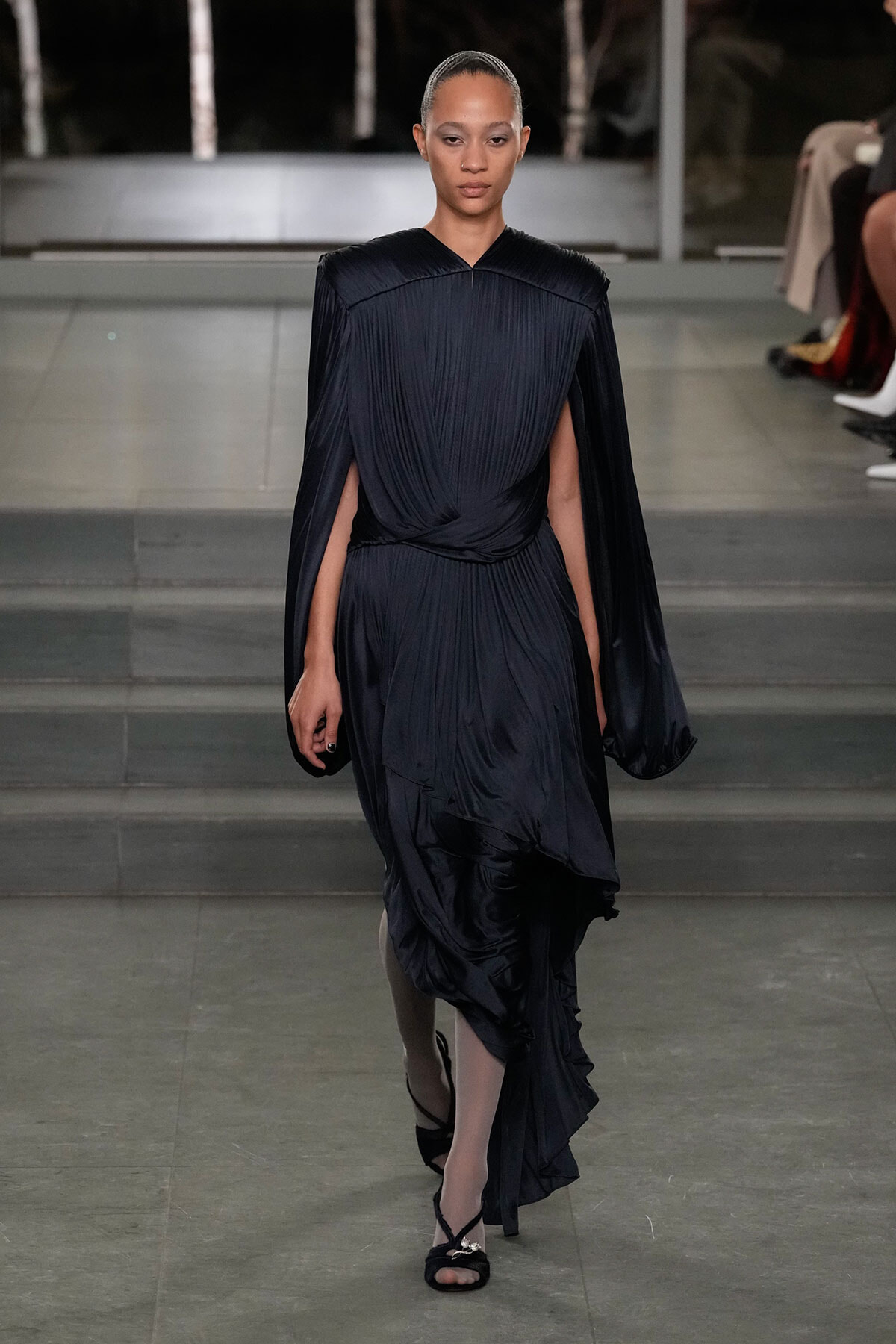 Model walks the runway in a dark navy pleated gown with cape sleeves and a draped, twisted waist.