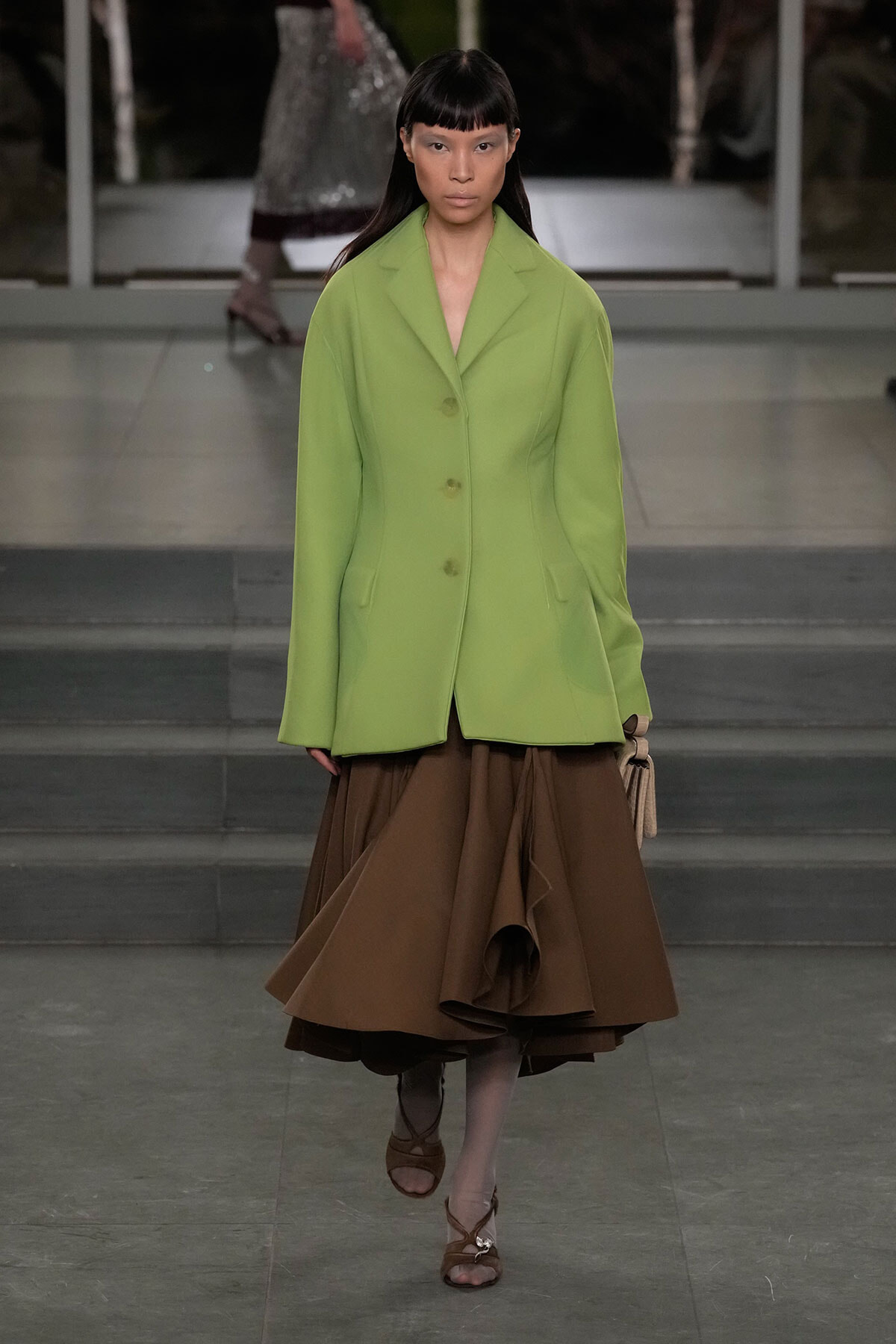 Model walks a runway in a lime green blazer paired with a voluminous brown skirt.