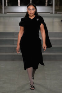 Model walking a runway in a black velvet wrap dress with a brooch on the shoulder, neutral makeup, and strappy black heels.