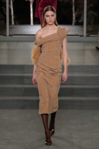 Runway model wearing a tan one-shoulder, ruched midi dress with fur-trimmed sleeves walking the catwalk.
