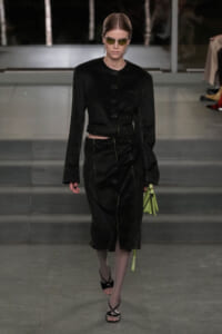 Model on a runway in a black cropped jacket and matching skirt with prominent buttons, wearing yellow-tinted sunglasses and carrying a lime-green handbag.