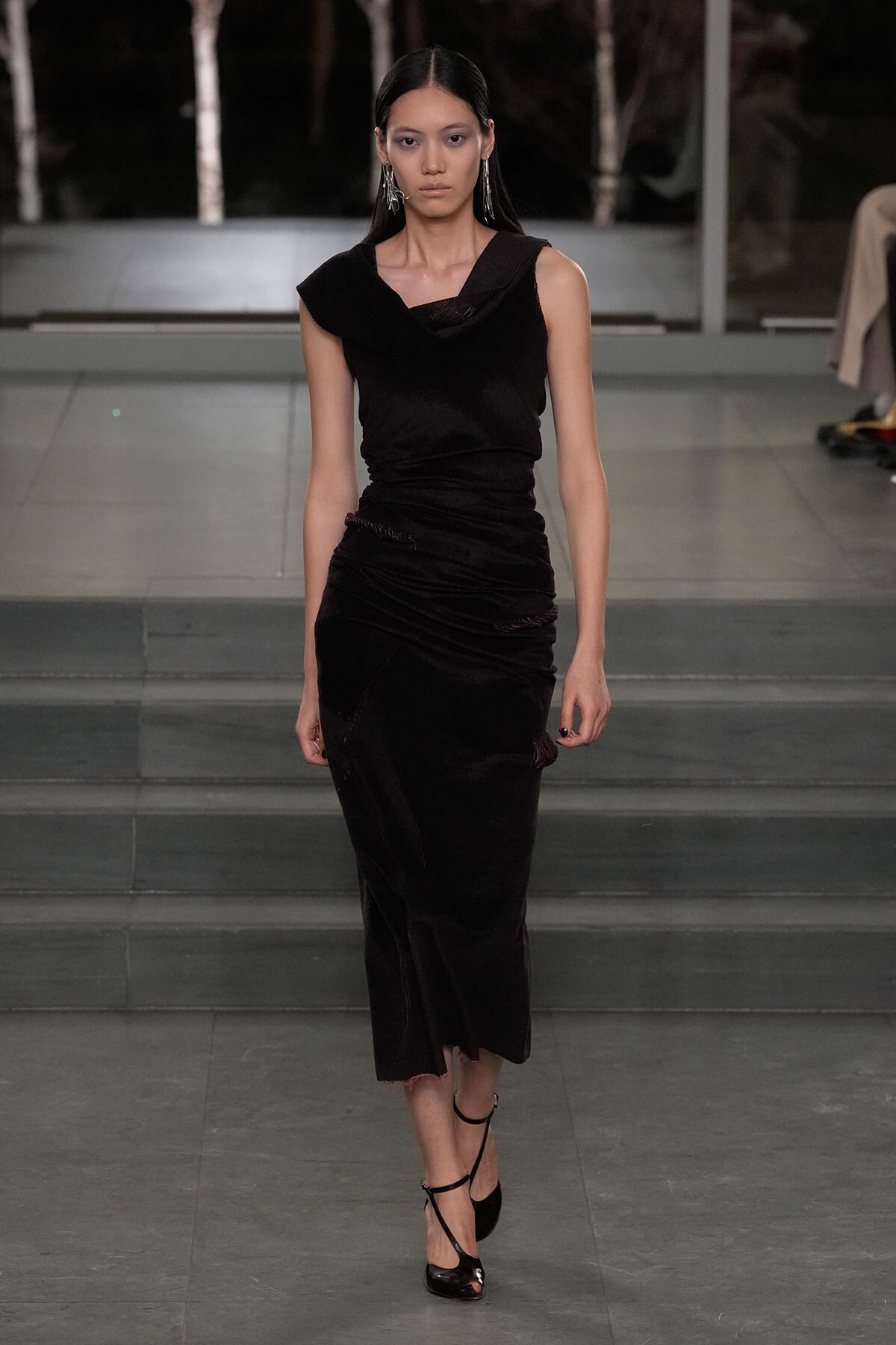 Model walking the runway in a sleeveless black draped dress with statement earrings and strappy heels.