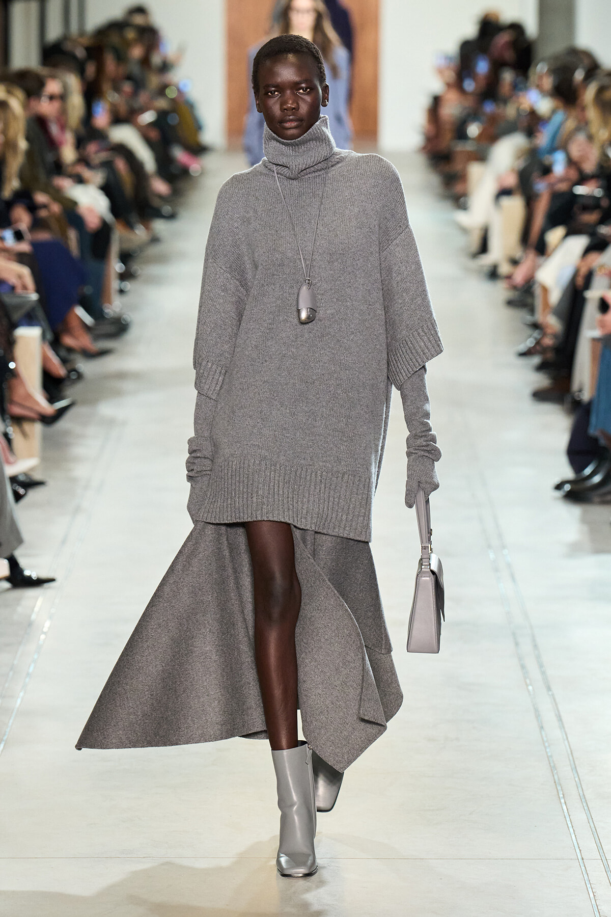 Runway model in a monochrome gray knit outfit: oversized turtleneck sweater dress over a flowing asymmetric gray skirt, with silver boots and a pendant necklace.