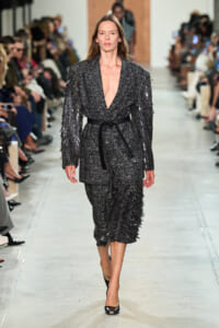 Model walking a runway in a charcoal textured blazer with fringe details and a matching fringe skirt, cinched with a black belt and black heels.