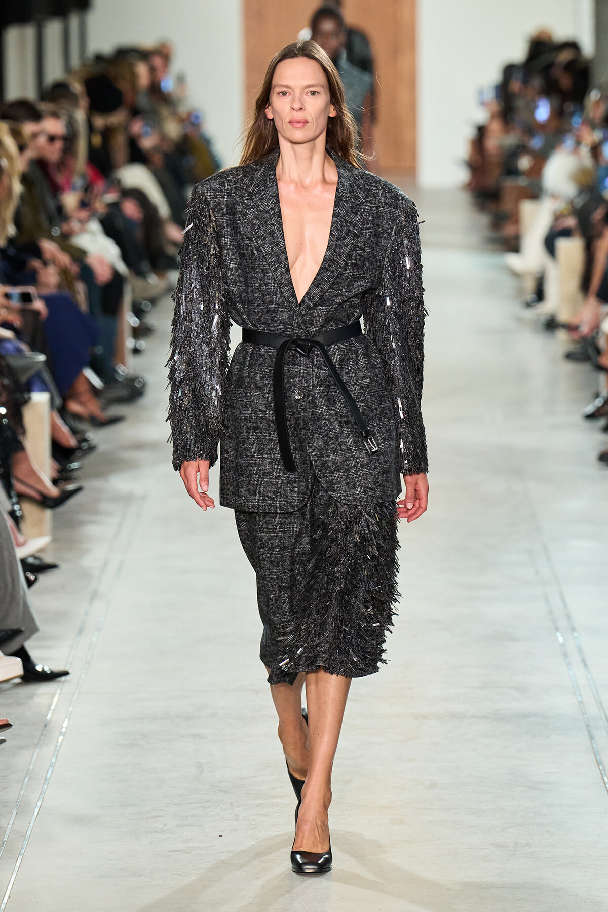 Model walking a runway in a charcoal textured blazer with fringe details and a matching fringe skirt, cinched with a black belt and black heels.