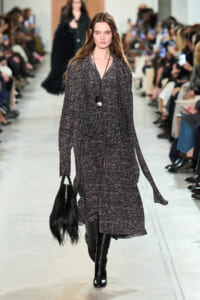 Model walks a textured gray coat down the runway, paired with black knee-high boots and a black fringe handbag; audience watches on both sides.