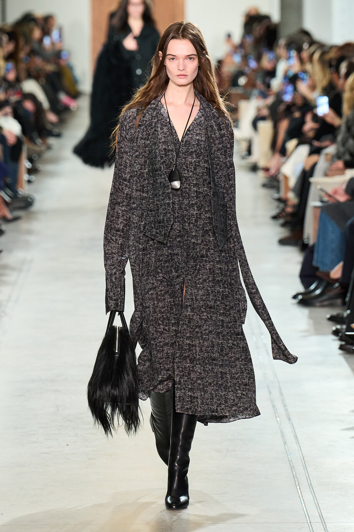 Model walks a textured gray coat down the runway, paired with black knee-high boots and a black fringe handbag; audience watches on both sides.