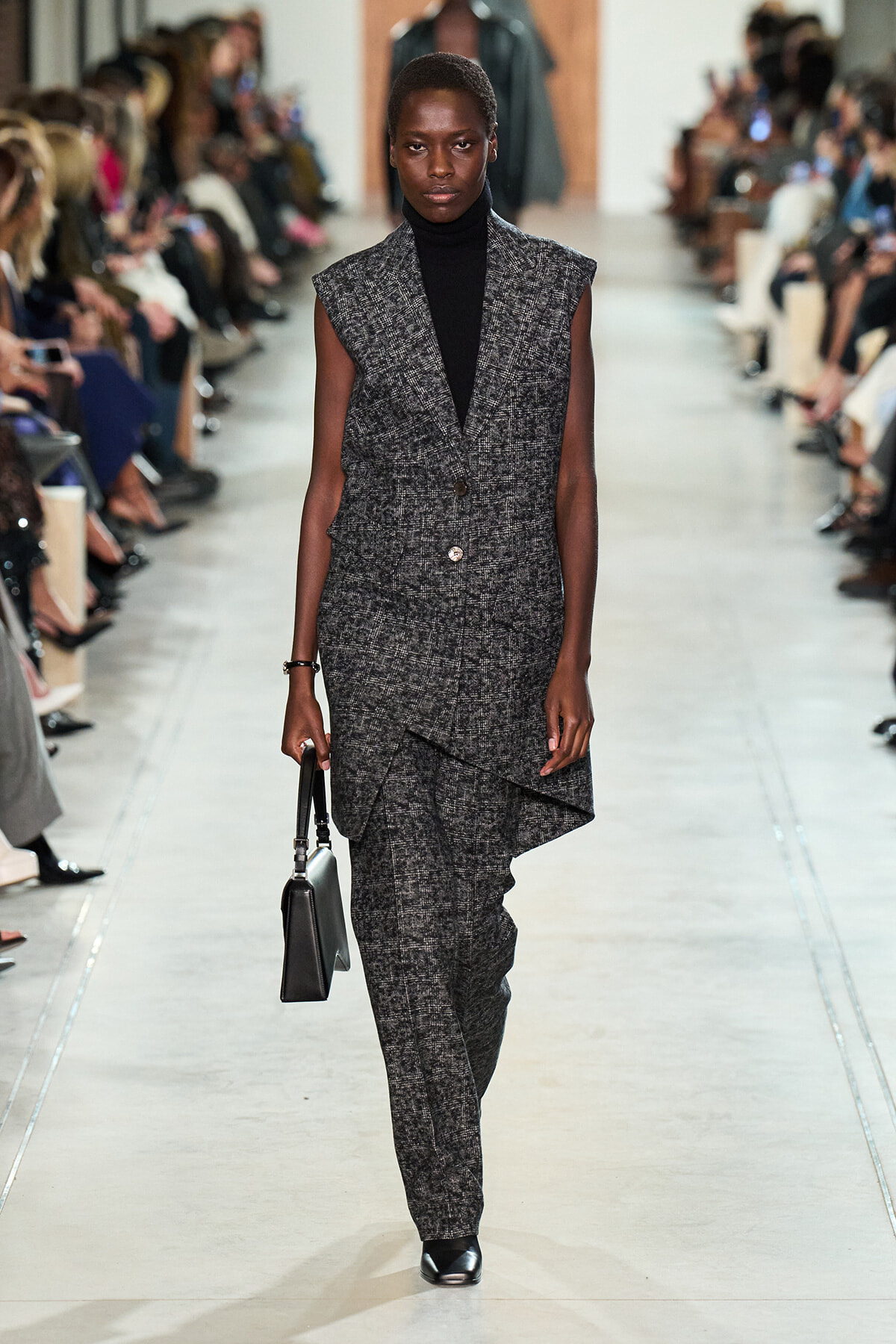 Model walking a fashion runway in a gray tweed sleeveless blazer with matching pants, black turtleneck, and a small black handbag.