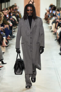 Male model walks a runway in a long gray overcoat, black turtleneck, gloves, and sunglasses, carrying a black fur-accented handbag.