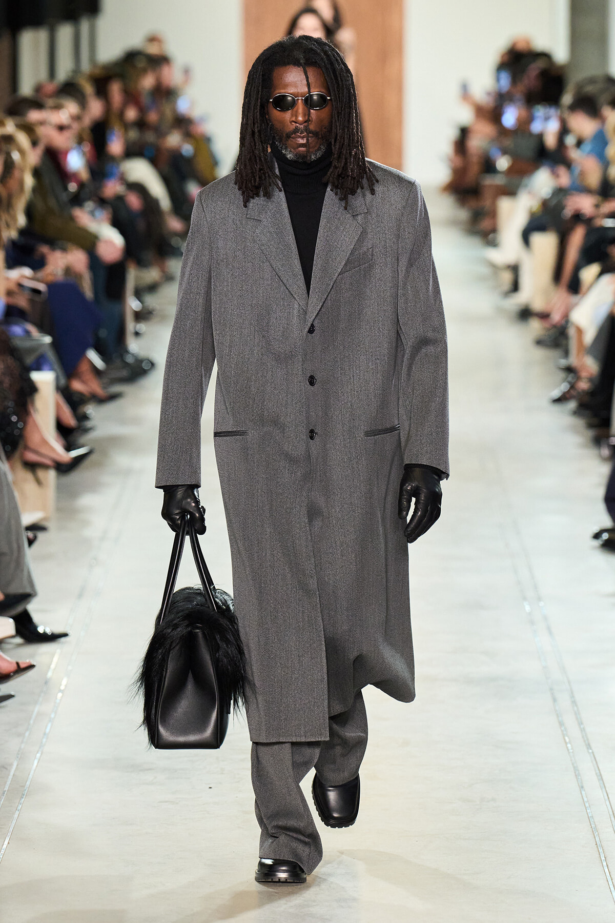 Male model walks a runway in a long gray overcoat, black turtleneck, gloves, and sunglasses, carrying a black fur-accented handbag.