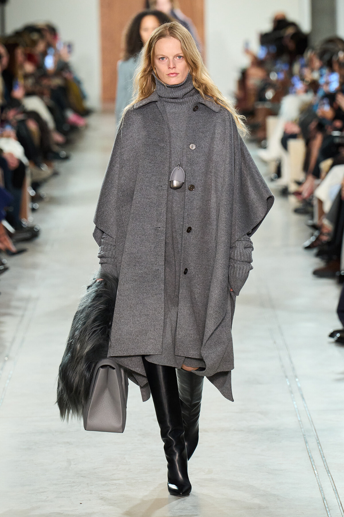 Model walks a fashion runway in a long gray cape coat, gray turtleneck, and black knee-high boots, carrying a gray handbag and faux fur stole on her left arm