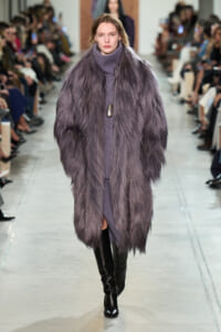 Model on a fashion runway wearing a long lavender fur coat over a matching turtleneck dress and black knee-high boots, with seated audience along the runway.