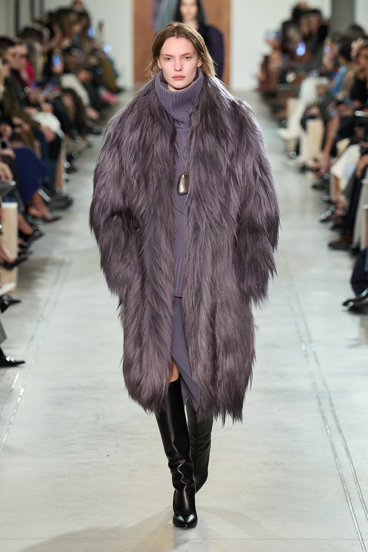 Model on a fashion runway wearing a long lavender fur coat over a matching turtleneck dress and black knee-high boots, with seated audience along the runway.