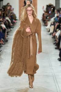 Model on a runway wearing a camel wrap coat with a belt, tan fur stole draped over her left arm, and knee-high tan boots.