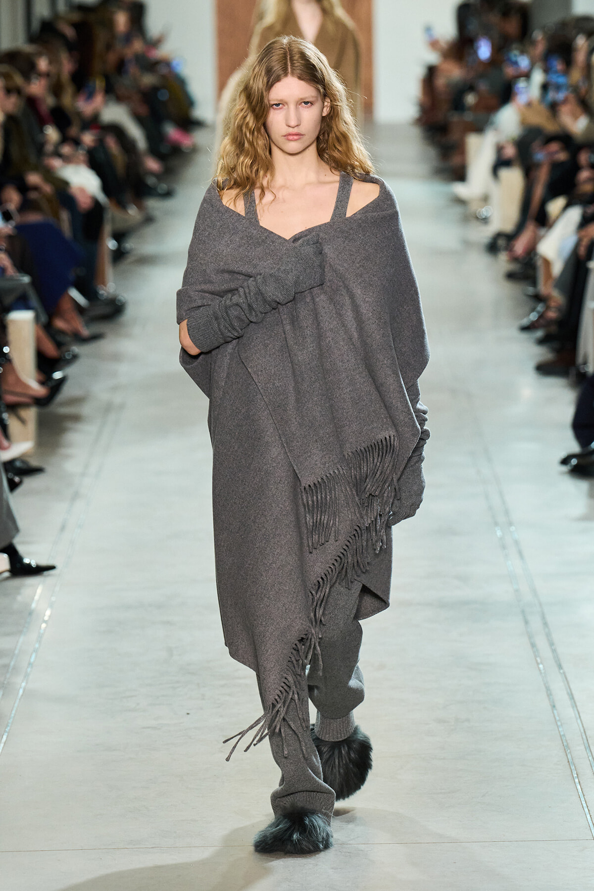 Model walking the runway in a loose gray poncho with fringe, pairing gray knit pants and furry slippers.