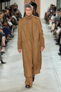 Male model walking a runway in a long camel overcoat and brown turtleneck, wearing round glasses, audience on both sides