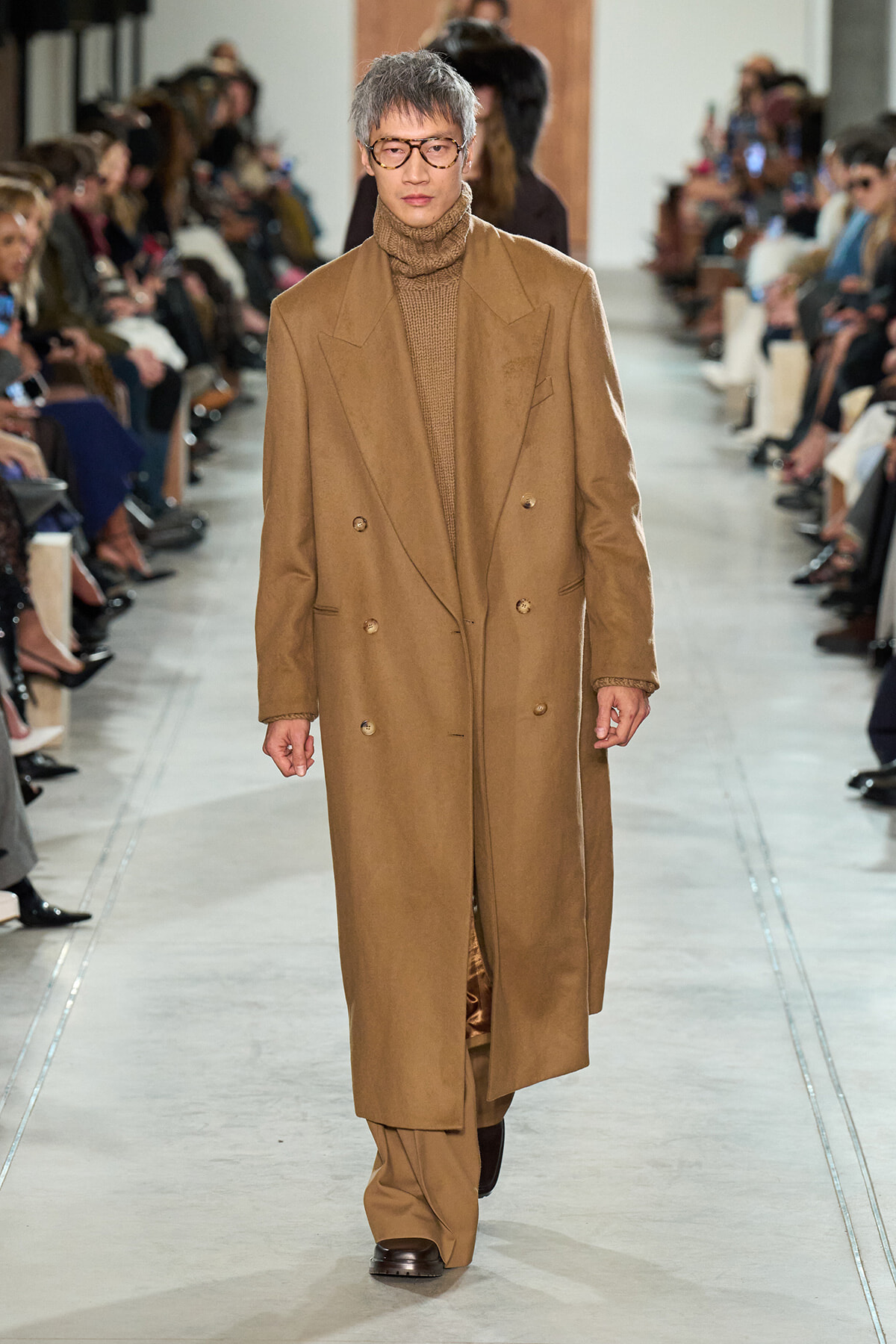Male model walking a runway in a long camel overcoat and brown turtleneck, wearing round glasses, audience on both sides