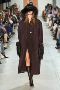 Model walking a runway in a dark brown long coat with oversized black fur cuffs and a large fur hood, paired with knee-high brown boots and a pendant necklace.